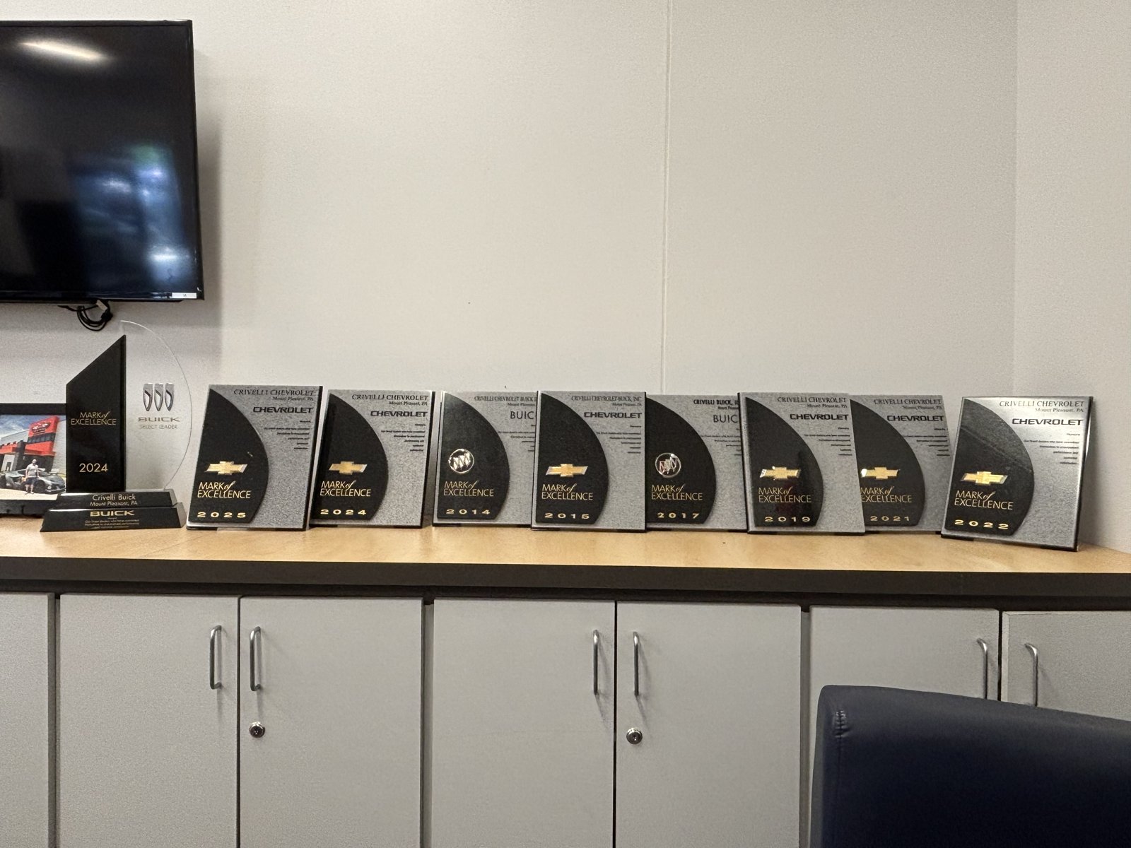 Crivelli Chevrolet Buick's shelf of GM Mark of Excellence award plaques for 2014, 2015, 2017, 2019, 2021, 2022, 2024, and 2025, in Mount Pleasant, PA